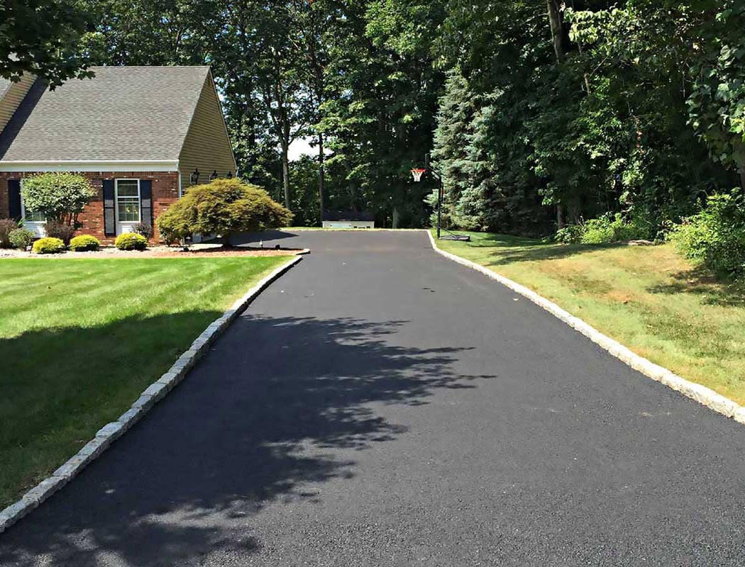 00005residential-1050×800-1 Freshly paved black asphalt driveway curves past a house with green lawn and trees.
