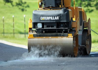 A steamroller compacts hot asphalt on a road, with steam rising from the fresh pavement.