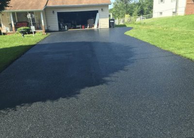 Freshly sealcoated driveway in front of a garage, with surrounding grass and houses visible.