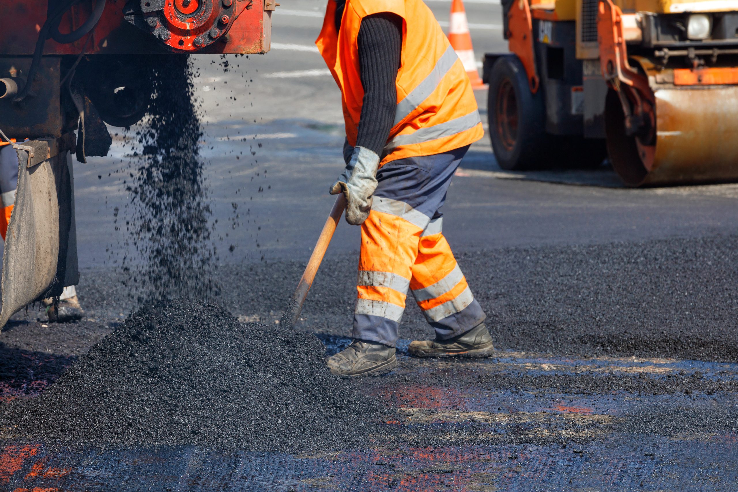 Worker in orange safety gear spreads fresh asphalt on a road using a shovel near paving machinery.