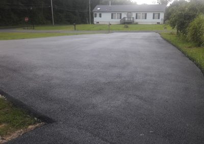 Freshly paved black asphalt driveway leading to a white house with trees in the background.