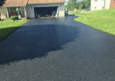 A driveway halfway paved with fresh black asphalt leads to a garage by a house with a lawn.