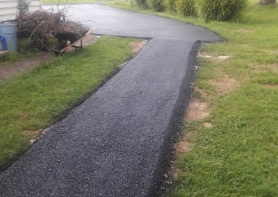 A newly paved asphalt pathway curves through a grassy yard beside a house on a cloudy day.