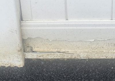Peeling white paint and damaged wood at the bottom of a white door above a black asphalt surface.