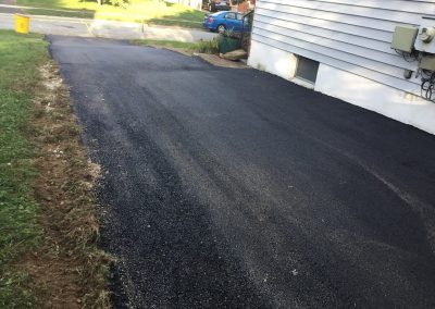 Freshly paved black asphalt driveway beside a white house, bordered by green grass and a curb.