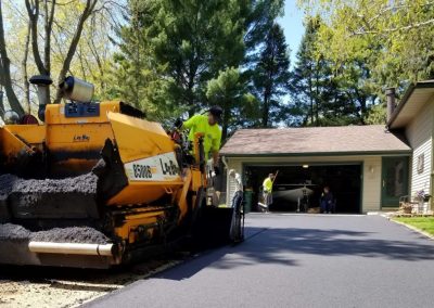 Workers paving a residential driveway with a yellow asphalt machine on a sunny day.