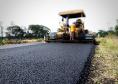 A road paving machine lays fresh asphalt on a rural road, with trees in the background.
