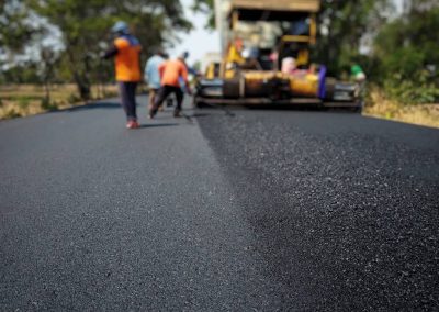 Workers paving a road with fresh asphalt, using machinery and tools, with trees in the background.