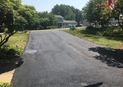 Freshly paved asphalt driveway leading to a house, surrounded by green trees and grass on a sunny day.