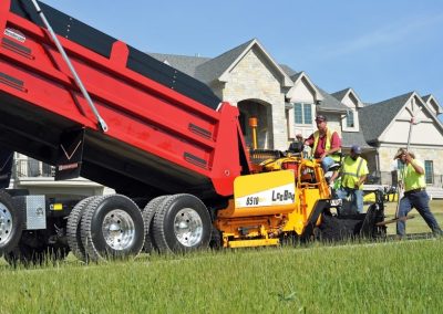 Workers operate paving equipment and a dump truck to lay asphalt near a suburban house on a sunny day.