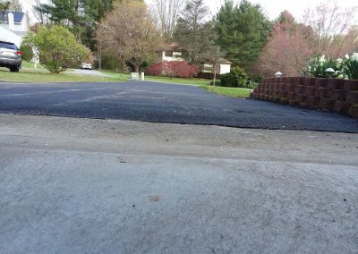 View of a driveway leading to a suburban street with trees, grass, and houses in the background.