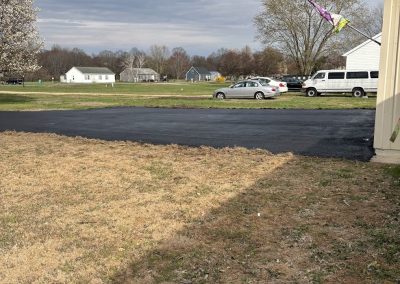 A patchy brown lawn beside a paved lot, cars and houses in the background under a cloudy sky.