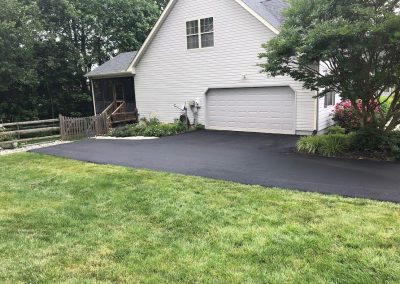 Freshly paved driveway leading to a white garage, with green lawn and trees surrounding the house.