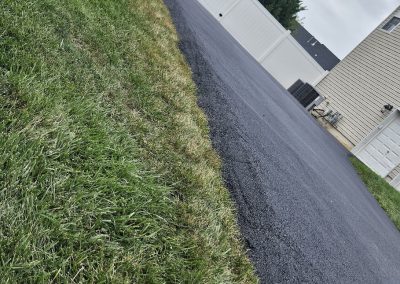 A driveway with fresh black asphalt runs beside a strip of green grass and a white fence in the background.