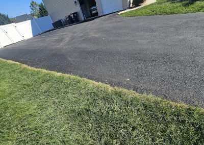 Freshly paved asphalt driveway beside a grassy lawn leading up to a garage on a sunny day.