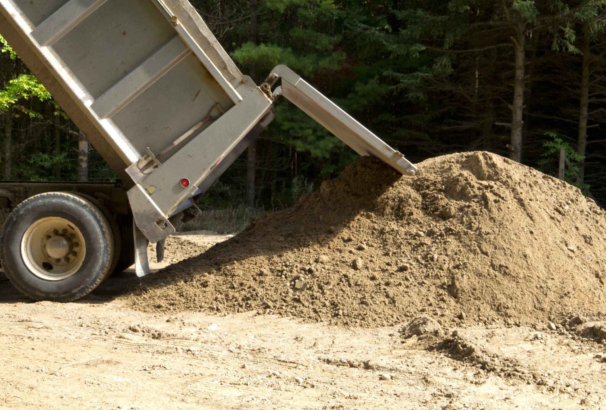 A dump truck unloading a large pile of dirt on the ground near a wooded area.
