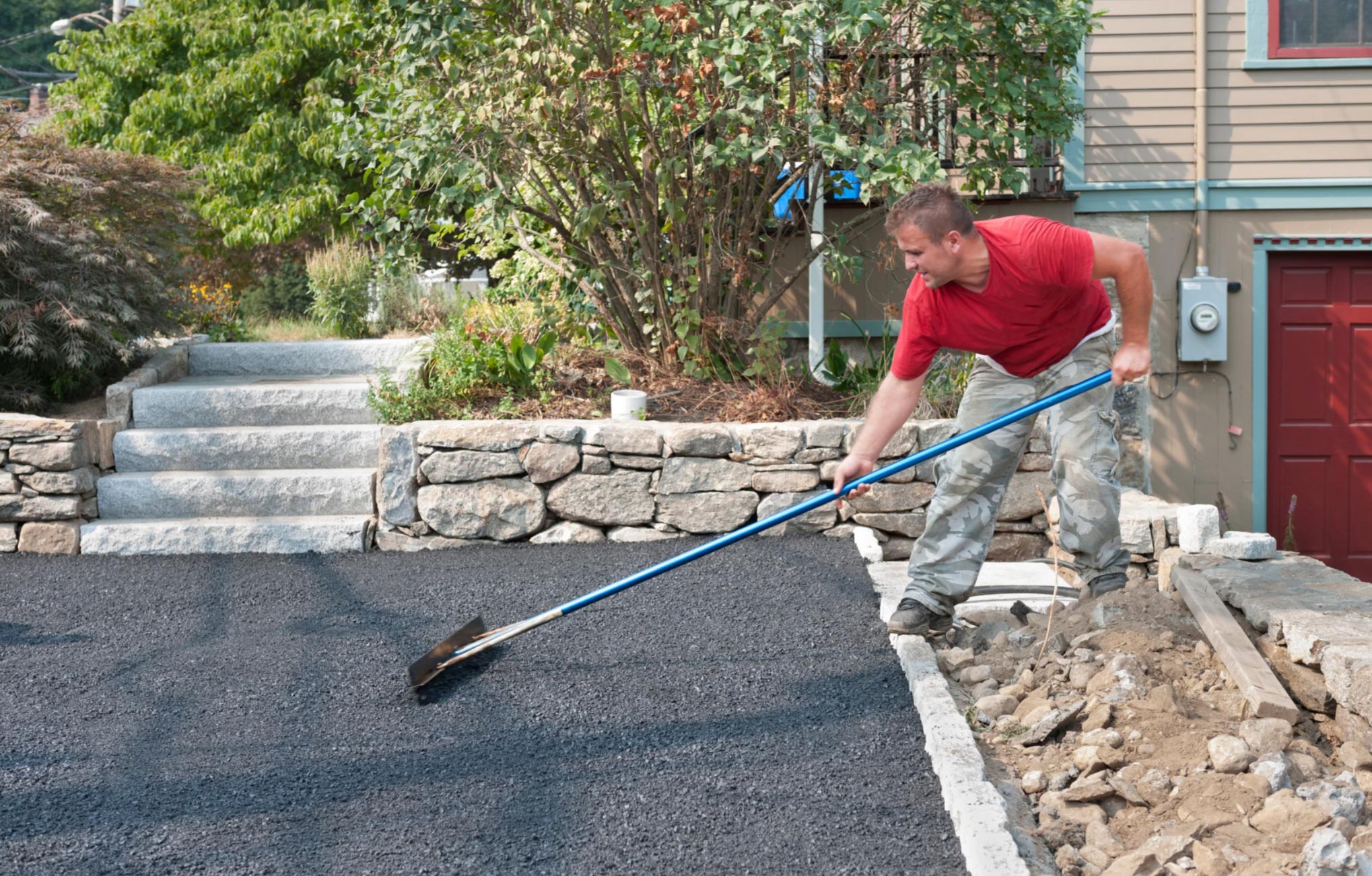 tar-chip-2 A man in a red shirt levels fresh asphalt on a driveway with a long-handled tool near a stone wall.