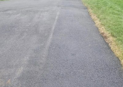 Freshly paved asphalt driveway next to green grass, leading to a street and trees in the background.
