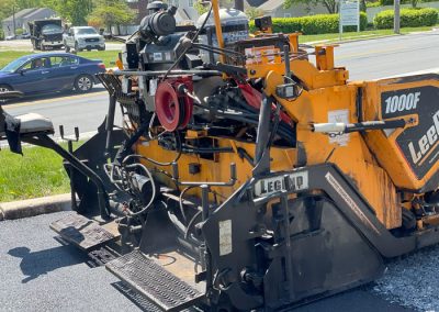 A yellow LeeBoy 1000F paving machine laying fresh asphalt on a sunny day near a road.