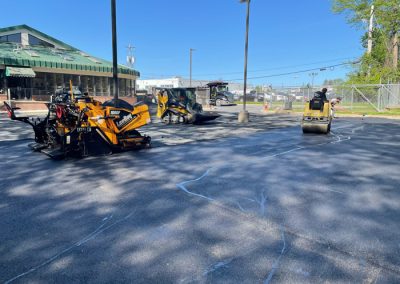 Workers paving a parking lot with construction equipment on a sunny day near a building and fence.