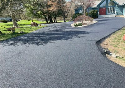 Freshly paved black asphalt driveway curving toward a blue house with red doors, surrounded by trees.