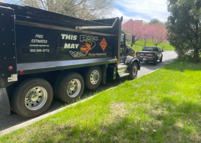 Black dump truck parked on a street, with grass and trees nearby on a sunny day.