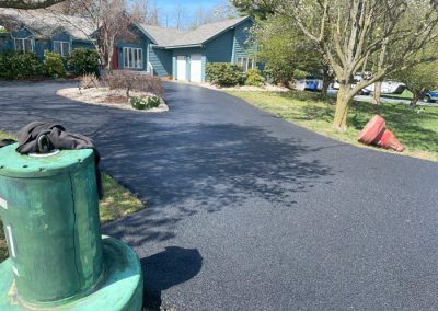 Freshly paved black asphalt driveway curves toward a blue house, with trees and utility equipment nearby.