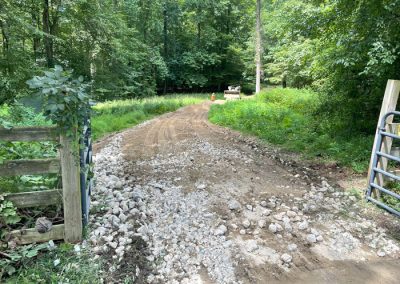 A dirt and gravel road curves through a lush green forest, with a wooden fence and gate on either side.
