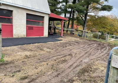 A barn with red trim, fresh asphalt, muddy tire tracks, and trees with autumn leaves in the background.