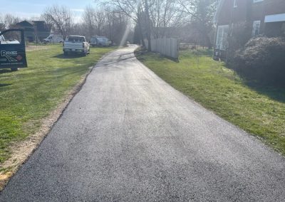 Freshly paved asphalt driveway on a sunny day, with grass and vehicles along the sides.