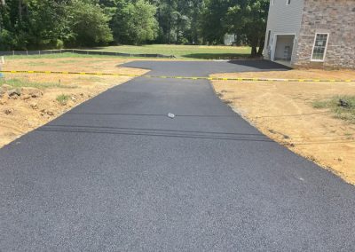 Freshly paved asphalt driveway blocked by caution tape, leading to a house with a stone exterior.