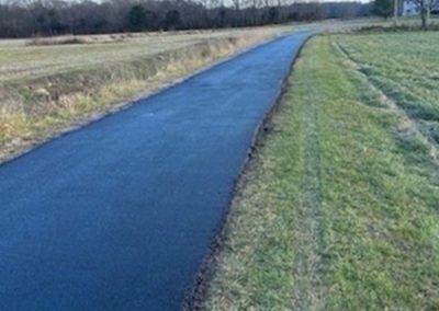 A newly paved path runs through open grassy fields with trees visible in the distance.