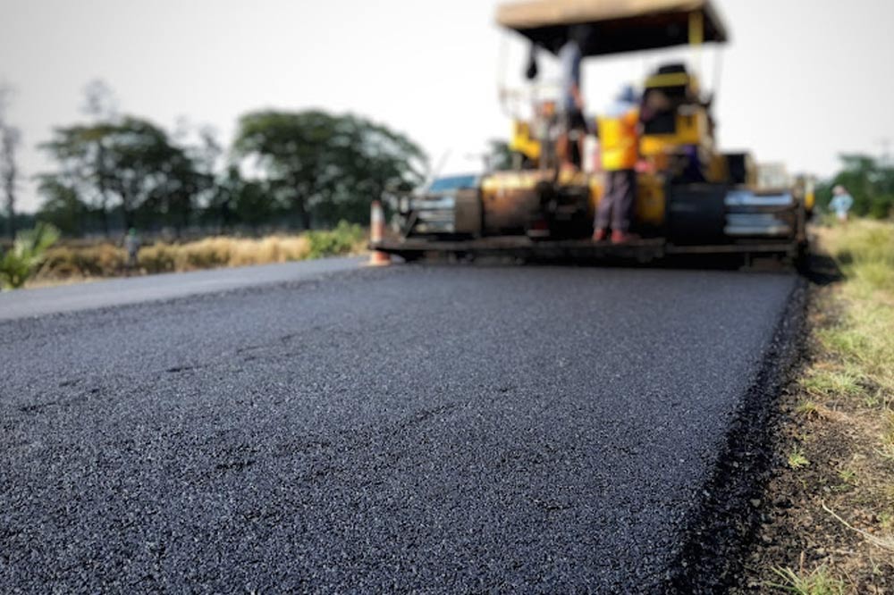 A close-up of fresh asphalt being laid on a road by a paving machine with workers.