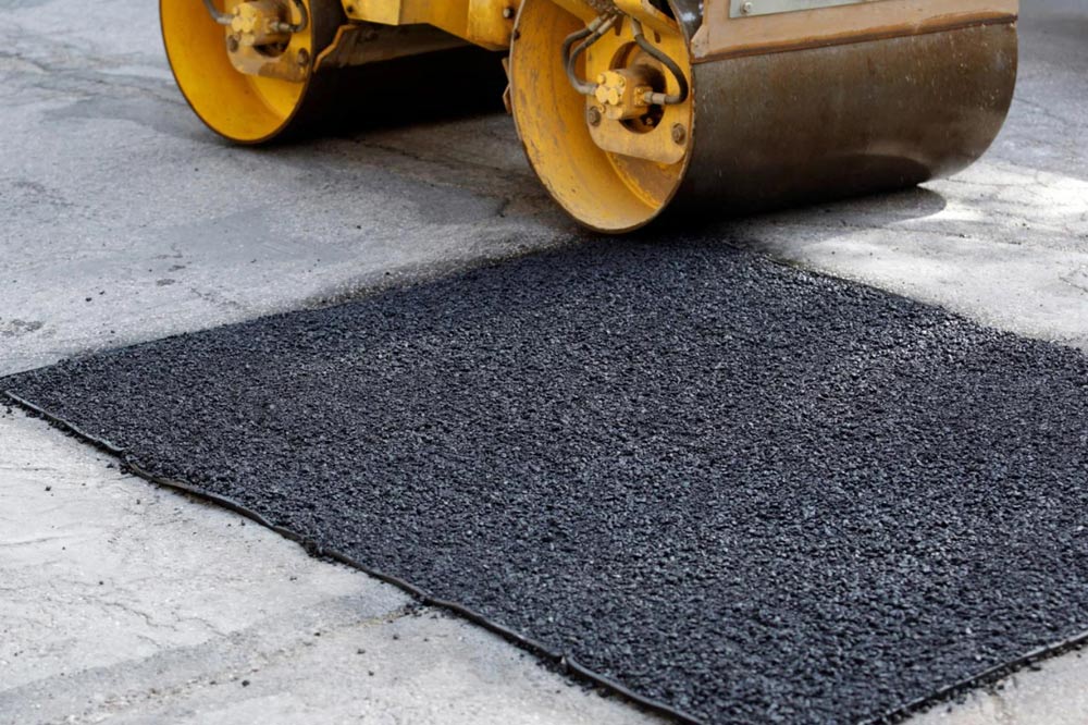 A road roller compresses fresh black asphalt patch on a gray concrete surface.