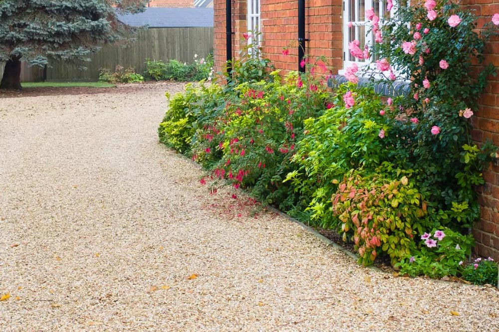 Gravel driveway beside a brick house with colorful flowers and green shrubs along the edge.