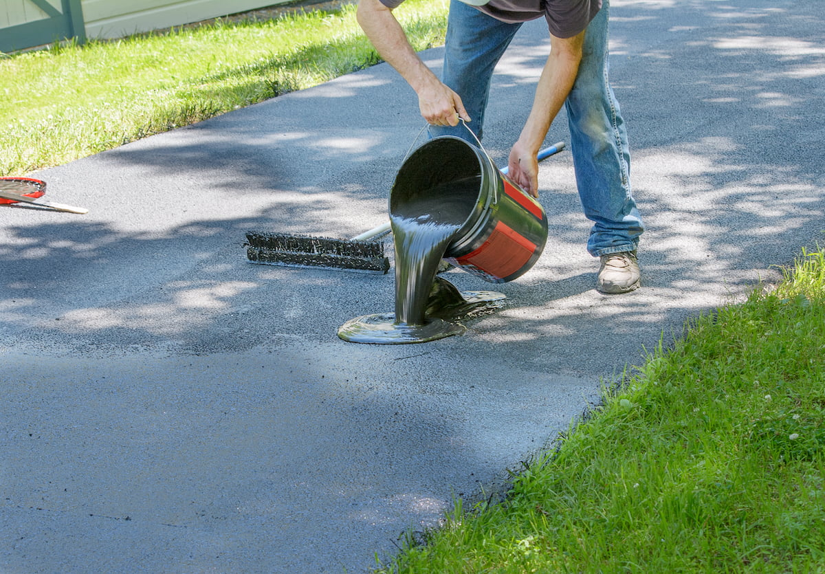 A person pours black asphalt sealer from a bucket onto a driveway in a grassy yard.